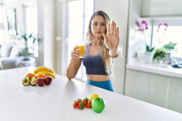 Young caucasian fitness woman wearing sportswear drinking healthy orange juice with open hand doing stop sign with serious and confident expression, defense gesture  - 写真・画像