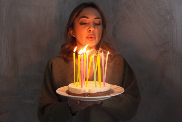 Mladá usmívající se žena drží domácí narozeninový dort s hořícími svíčkami, zatímco stojí uvnitř, boční pohled. Samice nosí koláč Bday. Narozeninové tradice a koncepce oslav - Fotografie, Obrázek