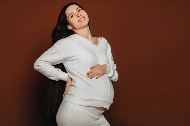 Молодая счастливая беременная женщина с длинными каштановыми волосами трогает живот, разговаривает с нерожденным ребенком, находясь в изоляции на темном студийном фоне, будущая мать в белой одежде гладит животик. Беременность - Фото, изображение