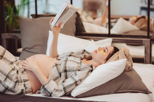 Junge attraktive, glückliche Schwangere in Homewear, die Buch liest, den Bauch berührt, während sie sich zu Hause auf der Couch entspannt, die werdende Mutter im karierten Pyjama die Freizeit während der Schwangerschaft genießt - Foto, Bild
