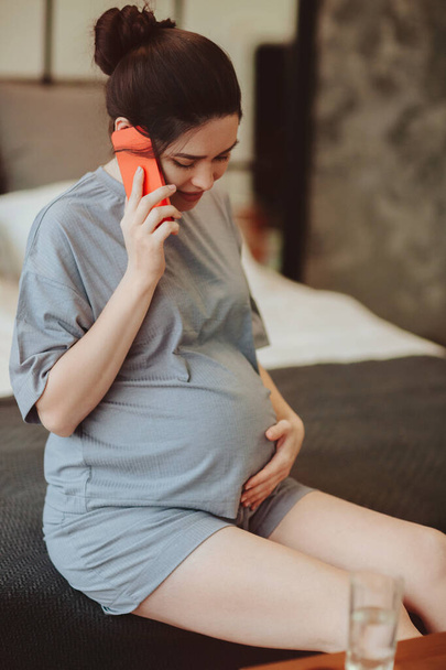 Junge lächelnde Schwangere, die den Bauch berührt, während sie zu Hause mit einem Freund telefoniert, werdende Mutter in Hauskleidung, die auf dem Bett sitzt, den Bauch streichelt und mit dem Smartphone telefoniert - Foto, Bild