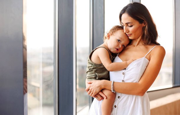 Bycie mamą. Młoda piękna kobieta matka trzyma słodkie dziecko chłopiec stojąc przy oknie w domu. Kobieta w białej sukience przytula swojego synka i wyraża miłość i troskę - Zdjęcie, obraz