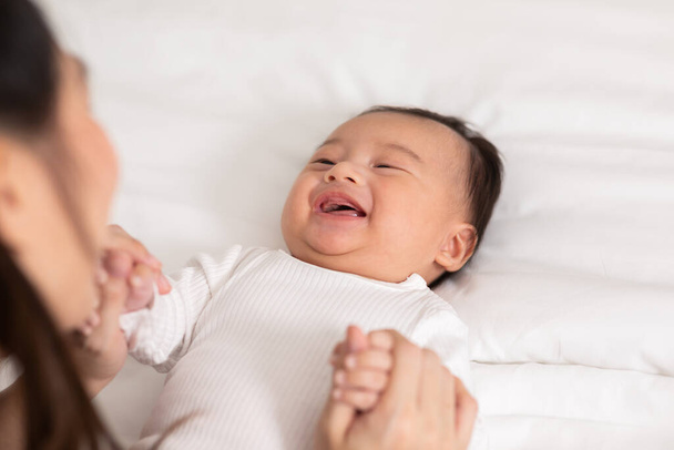 New asian mom playing to adorable newborn baby on bed smiling and happiness at home.Mom talking with infant baby and holding her hands laughing together.Baby and Mother day concept - Фото, изображение