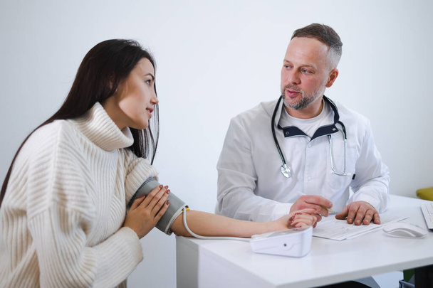 A férfi orvos méri a nő vérnyomását, és rögzíti az eredményeket a beteg kártyáján. Nő a kardiológusnál - Fotó, kép