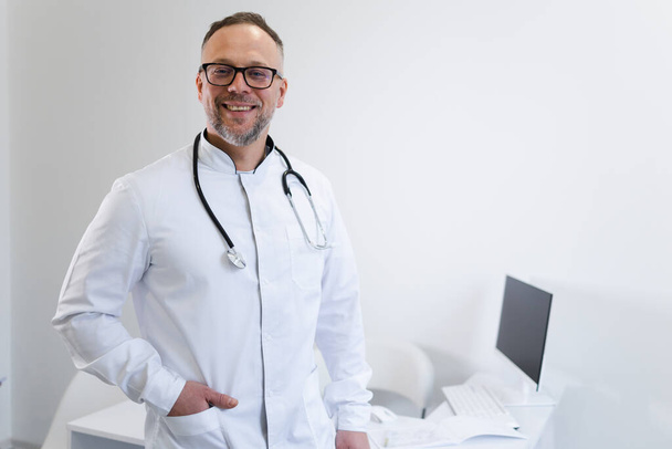 Glimlachende mannelijke arts met stethoscoop om zijn nek kijkend naar de camera  - Foto, afbeelding
