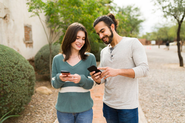 novio latino y novia caucásica sonriendo mientras los mensajes de texto en sus teléfonos. Hombre guapo mostrando una foto en su teléfono inteligente  - Foto, Imagen