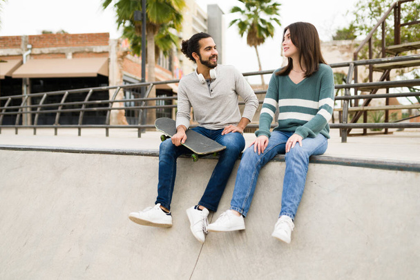 Лучшие друзья смешанной расы улыбаются, тусуясь в скейт-парке и разговаривая, держа в руках скейтборд - Фото, изображение