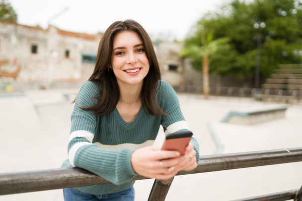 Mulher bonita em seus 20 anos olhando para a câmera e sorrindo enquanto segurava seu smartphone ao ar livre  - Foto, Imagem