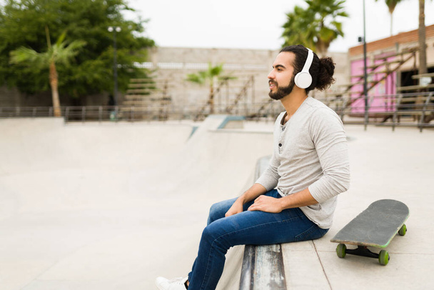 Вид збоку розслабленого молодого чоловіка, який слухає музику з бездротовими навушниками та скейтбордингом у парку
 - Фото, зображення