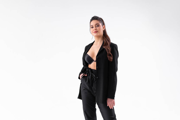Young business woman with stretched black jacket from under which black bra is visible standing studio isolated on white background. Caucasian female business person looking at cameara. Medium shoot - Zdjęcie, obraz