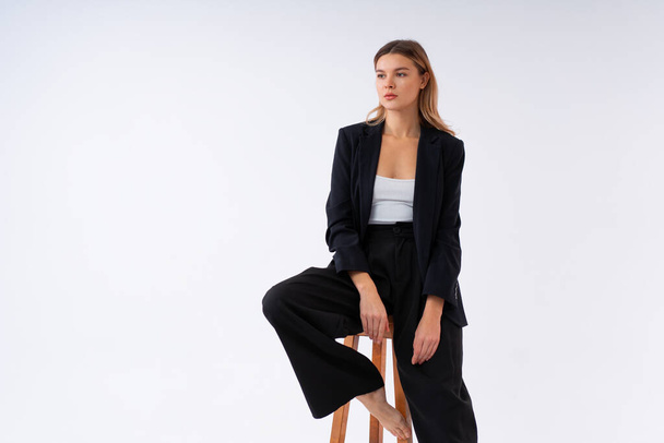 Young fashionable business woman sitting high bar wooden chair dressed black office suit isolated in white background. Caucasian female business person pensive looking away with bare foot - Fotó, kép