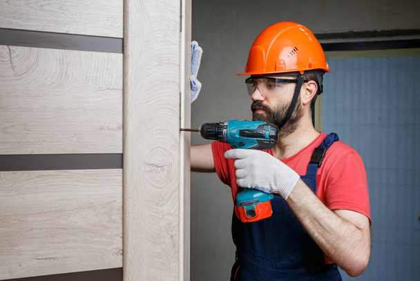 Turuncu kasklı matkaplı bir işçi eve bir kapı yerleştirir. - Fotoğraf, Görsel