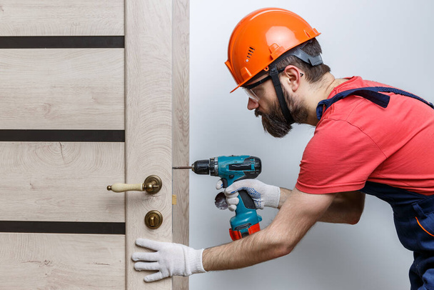 un trabajador con un taladro en un casco naranja instala una puerta en la casa - Foto, imagen