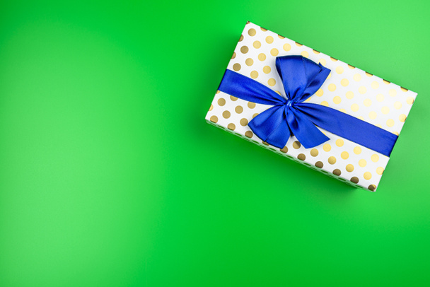 un cadeau enveloppé dans du papier blanc avec des cercles d'or enveloppés dans un ruban bleu attaché dans un arc, isolé sur un fond vert, vue du dessus. - Photo, image