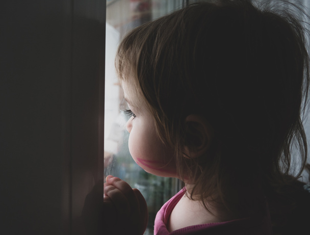 een klein meisje is verdrietig en kijkt uit het raam, ze wil door de straat lopen. Het kind kijkt uit het raam. Portret van een droevige baby op de vensterbank. - Foto, afbeelding