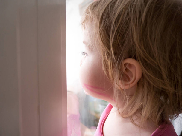 une petite fille est triste et regarde par la fenêtre, elle veut marcher dans la rue. L'enfant regarde par la fenêtre. Portrait d'un bébé triste sur le rebord de la fenêtre. - Photo, image