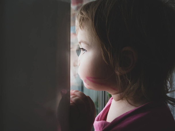 a little girl is sad and looks out the window, she wants to walk down the street. The child looks out the window. Portrait of a sad baby on the windowsill. - Zdjęcie, obraz