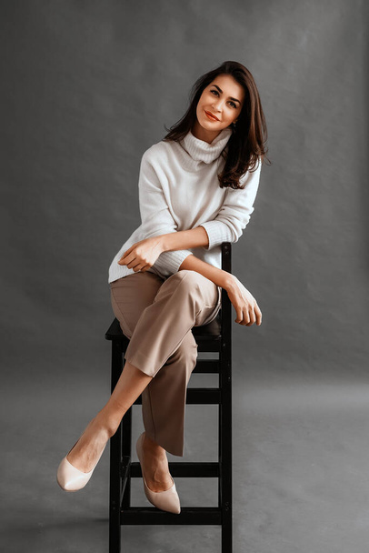 Zorgeloos jonge zakenvrouw zittend op een stoel, gekleed in witte trui en beige broek, poserend in studio over grijze achtergrond. Ontspannen en mooie jonge vrouw - Foto, afbeelding