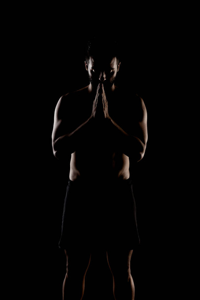 Side lit muscular Caucasian man silhouette. Athlete praying against black background.  - Foto, Bild