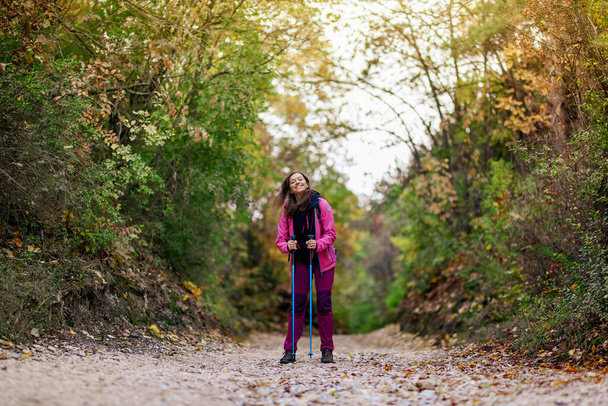 Hiker girl standing on a wide trail in the mountains. Backpacker with pink jacket in a forest. Healthy fitness lifestyle outdoors. - Photo, image
