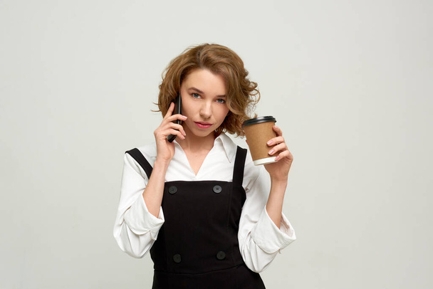Katı iş kadını telefonda konuşur ve gri arka planda kahve içer. - Fotoğraf, Görsel