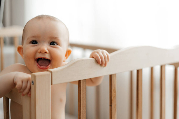 Portrait of a cheerful happy baby standing in the crib. The girl is standing alone in the bed. Copy space for text - Foto, afbeelding