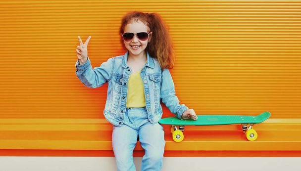 Portrait of cheerful laughing little girl child posing with skateboard in the city on an orange background - Fotó, kép