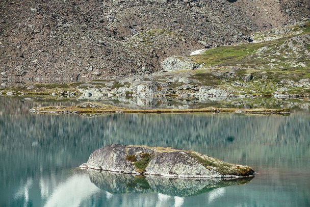 Rock in turquoise mountain lake. Snowy mountain reflected in azure clear water of glacial lake. Beautiful sunny background with snow-white glacier reflection in green water surface of mountain lake. - Photo, Image