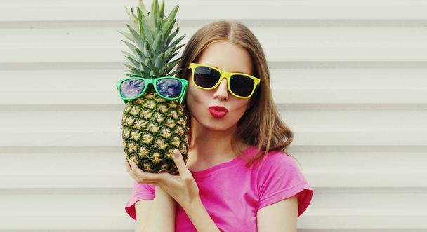 Summer portrait of young woman with pineapple blowing lips wearing a sunglasses over a white background - Photo, image