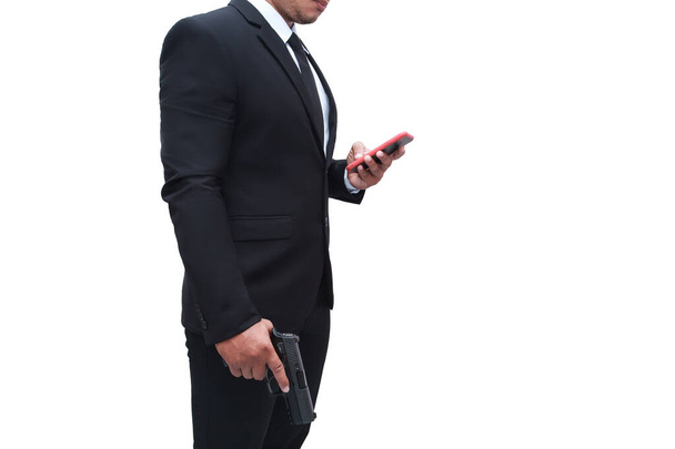 Ein bewaffneter Mann im schwarzen Anzug hält eine Pistole in der Hand und seine andere Hand geht zu seinem Handy, um sein Ziel auf weißem Hintergrund zu überprüfen. Konzept für Mörder, Mord, Verbrecher, Dieb. - Foto, Bild