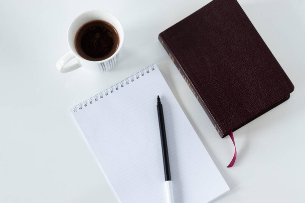 A blank notebook page with a closed Holy Bible Book, and a cup of coffee on white background with copy space for text. Top angle view. The biblical concept of Bible study and prayer. - Photo, Image