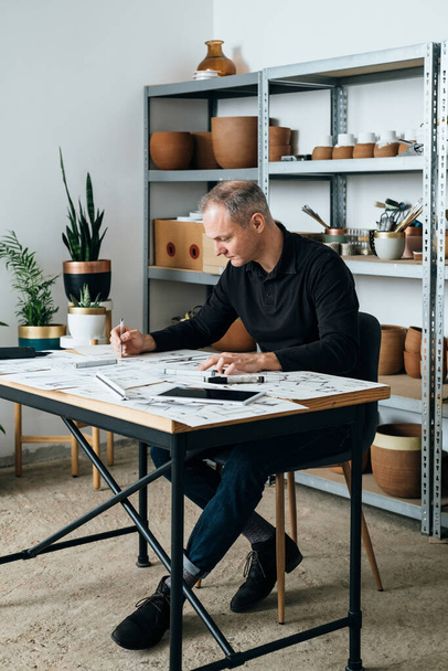 Serious Architect Working on an Architectural Drawing for Home Renovation. Sustainable Lifestyles: Successful designer sitting at the table and sketching blueprints at his workplace. - Photo, Image