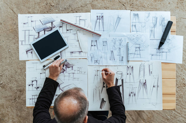 Ein anonymer Designer zeichnet Blueprint Design im Design Studio. Overhead-Foto eines unkenntlichen Möbeldesigners, der Zeichnungen mit Stift, Lineal und digitalem Tablet auf seinem Schreibtisch skizziert. - Foto, Bild