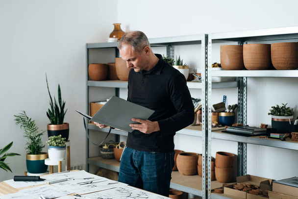 Serious Furniture Designer Analyzing Blueprints, Documents and Sketches in his Office. Successful architect or engineer holding folder and looking at his plan project while standing at his desk.  - 写真・画像