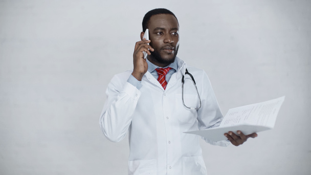 Африканский американский врач в белом халате разговаривает по мобильному телефону, держа папку с документами - Фото, изображение