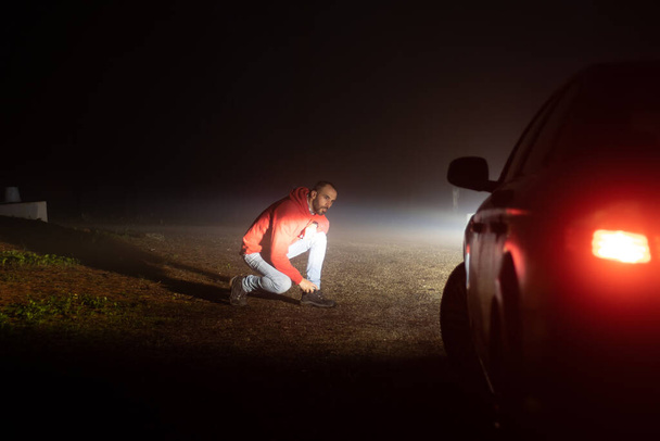 Hombre mirando a su coche roto en una carretera una noche brumosa, al aire libre - Foto, Imagen