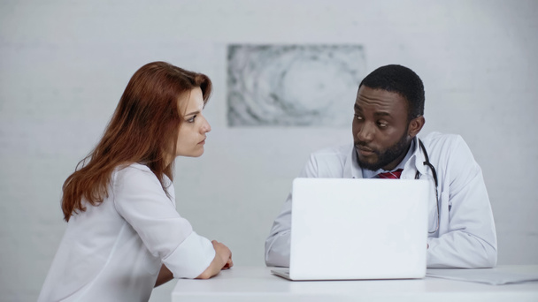 アフリカ系アメリカ人の医者がノートパソコンの近くでレッドヘッドの患者と話していて - 写真・画像