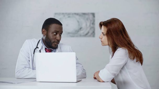 bearded african american doctor talking with redhead patient near laptop on desk - Foto, Imagen