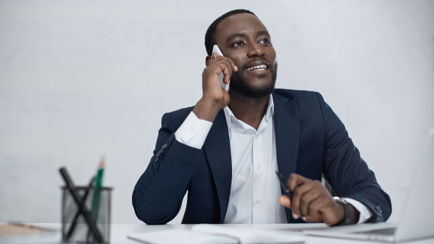happy african american businessman talking on smartphone near laptop on desk isolated on grey - Fotó, kép