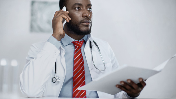 médico afroamericano hablando por teléfono móvil mientras sostiene la carpeta borrosa  - Foto, imagen