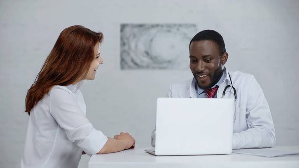 happy african american doctor talking with redhead patient near laptop on table  - Foto, imagen