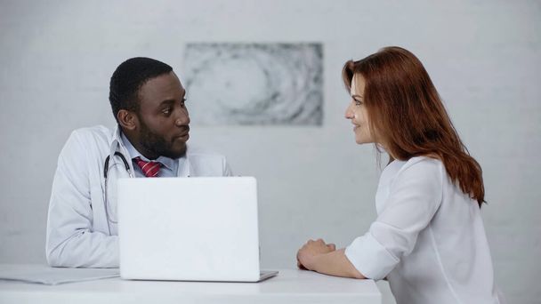 african american doctor talking with pleased redhead patient near laptop on table  - Zdjęcie, obraz