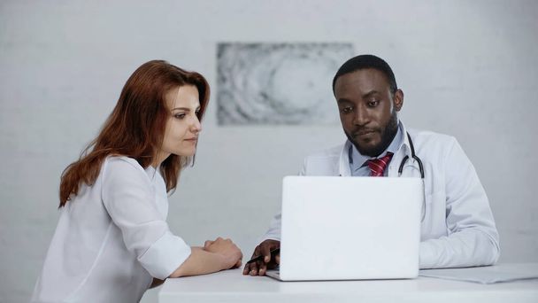 afro-amerikai orvos és vörös hajú beteg laptopot néz az asztalon  - Fotó, kép