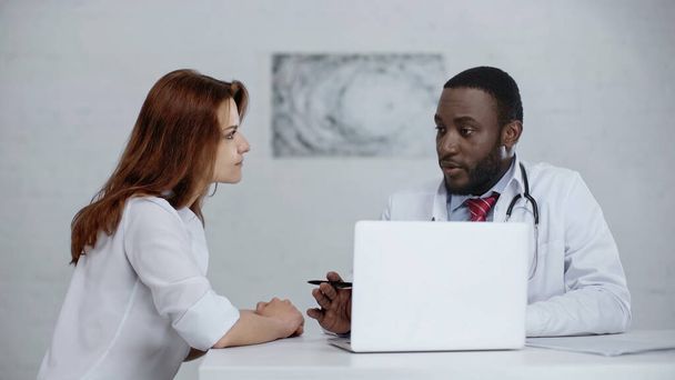 afro-amerikai orvos beszél vörös hajú nő mellett laptop az asztalon  - Fotó, kép