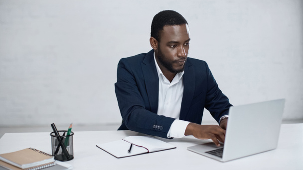 serious african american businessman typing on laptop near notebook on grey  - Fotoğraf, Görsel