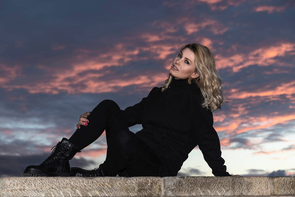 portrait of blonde woman dressed in black at sunset sitting on a wall - Фото, изображение