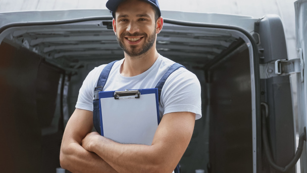 Cheerful delivery man crossing arms and holding clipboard near car outdoors  - Foto, imagen