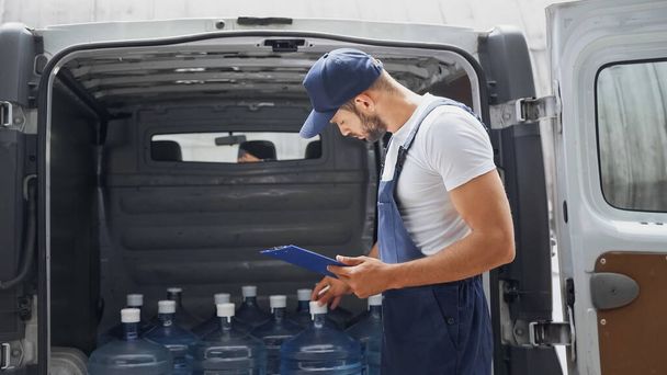 Side view of deliveryman in overalls holding clipboard near bottles of water in car  - Φωτογραφία, εικόνα