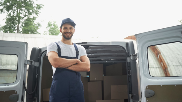 Smiling courier in cap and overalls looking at camera near boxes in car outdoors  - Photo, Image