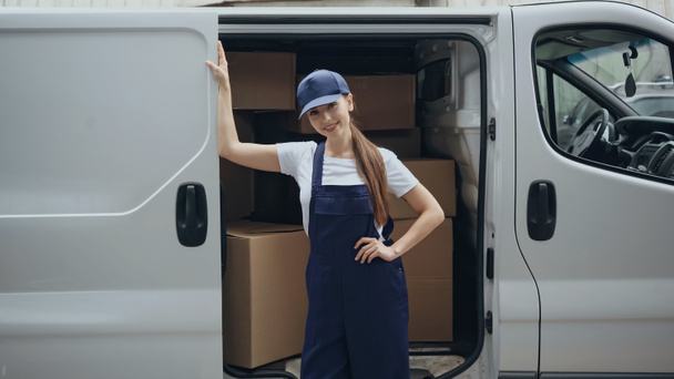 Smiling courier in overalls standing near cardboard boxes in car outdoors  - Photo, Image